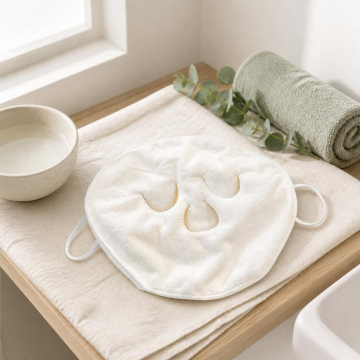 Compress face towel lying on a linen cloth on an oak bathroom shelf with a ceramic bowl, sage towel and eucalyptus