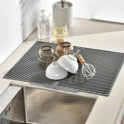 Grey silicone drying mat by the sink with glasses, a bowl and a whisk drying
