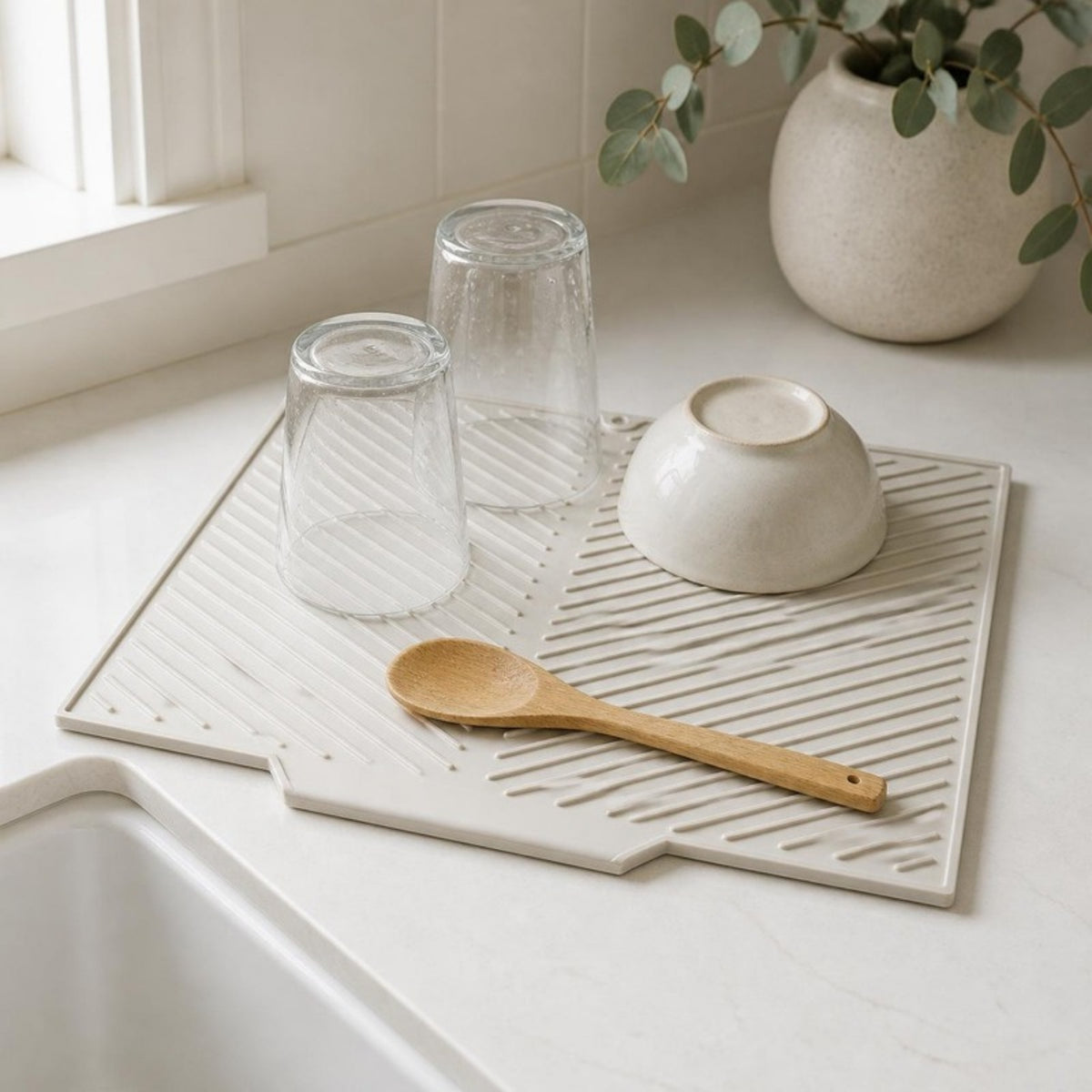 Light gray silicone drying mat beside a sink with upturned glasses, a bowl and a wooden spoon