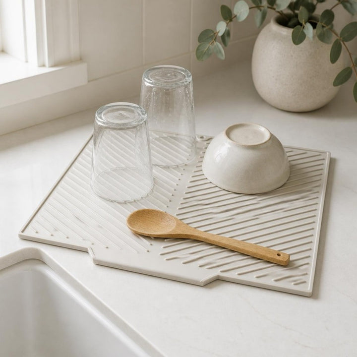 Light gray silicone drying mat beside a sink with upturned glasses, a bowl and a wooden spoon