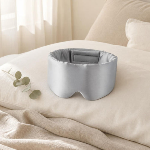 Light gray silk sleep mask resting on an ivory linen pillow in soft morning light