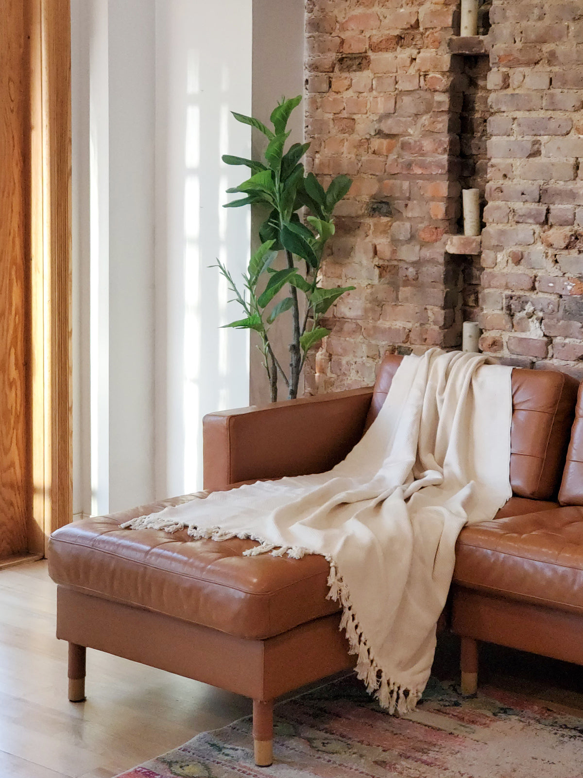 Artisanal cotton throw blanket with tassels draped over a tan leather sofa beside an exposed brick wall