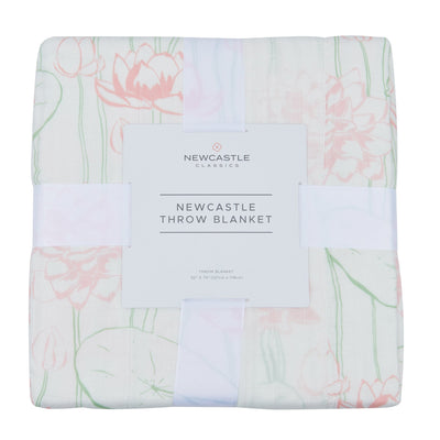A folded throw blanket with a pink and green floral pattern on a white background, wrapped in a white ribbon with a white paper label that reads 'NEWCASTLE CLASSICS NEWCASTLE THROW BLANKET'.
